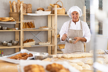 Professional aged female baker in white uniform and striped apron using whisk to mix ingredients in metal bowl, standing in small artisan bakery, surrounded by variety of freshly baked bread and tools