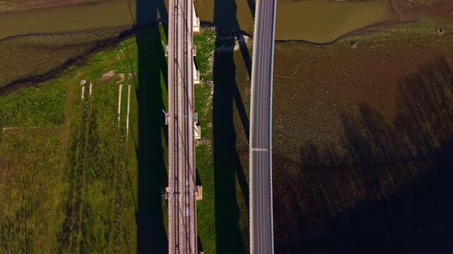 Railway tracks run parallel to a road bridge over a body of water in a rural setting. The shadows of the bridges stretch onto the ground below.