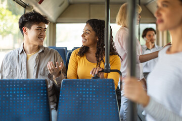 Portrait of happy cheerful couple sitting on city bus seat, young black lady and asian guy talking and looking at each other, enjoying ride or travel together in crowded public vehicle © Prostock-studio