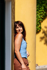 Fototapeta premium Young woman with brown hair is leaning against a yellow wall, showcasing a relaxed pose in a vibrant outdoor setting, capturing a moment of tranquility and warmth