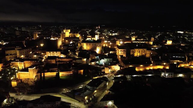 A 4K aerial night video filmed over the illuminated old town of Caceres in Extremadura, Spain. The drone moves slowly sideways, revealing the beautifully lit medieval historic center and stone