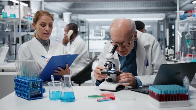 Laboratory technician examining biopsy slide while colleague documents diagnostic findings on clipboard. Elderly man identifies disease markers in clinical lab with coworker taking notes, camera B