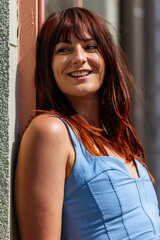 Fototapeta premium Smiling woman with long brown hair, wearing a light blue dress, leans against a wall in bright sunlight, showcasing a cheerful expression and relaxed atmosphere