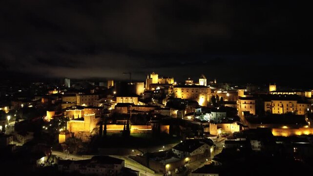 A 4K aerial night video filmed in the historic center of Caceres, Extremadura, Spain. The drone ascends slowly from a lower perspective, revealing the beautifully illuminated medieval old town at