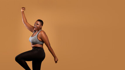 Obraz premium Excited black chubby woman in sportswear dancing, emotional happy lady celebrating success and victory, jumping, cheering and raising clenched fist up, brown studio background, banner, free copy space