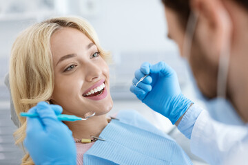 Closeup of male dentist in face mask doing treatment for patient cheerful young blonde lady,...