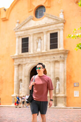 Fototapeta premium Young happy Latino man relaxed attitude walks near a historic colonial church in Cartagena, under bright sunlight, dressed casually in dark shorts, a red shirt, a shoulder bag and mirrored sunglasses