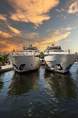 Fototapeta premium Two luxury yachts sit side by side, docked at a marina in Fort Lauderdale, Florida, as warm evening clouds drift across the sky in the background. The calm water reflects the vessels and soft sunset t