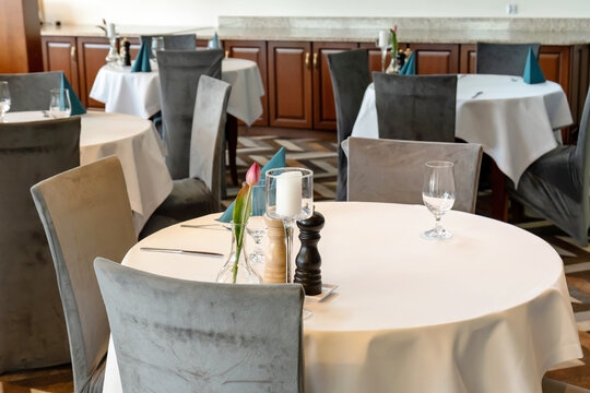 Empty dining room ready for event or booking. Elegant restaurant interior with round tables and grey chairs set for meal with white tablecloth, glassware, cutlery, napkins, candles and tulip flowers.
