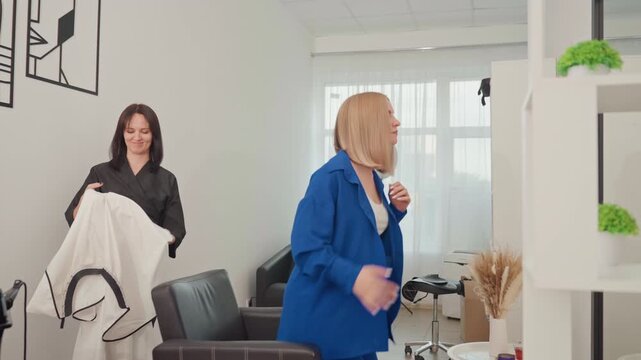 blonde caucasian client startled by cape, stylist holds white salon drape near modern chair while client on phone reacts with surprised laugh bright natural light, potted plants, minimalist shelving,