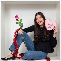 Young woman in a white box holding a rose and a pink heart sign with 'Feliz D&iacute;a'
