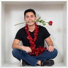 Young man with red rose in mouth and festive garland sits in a white box