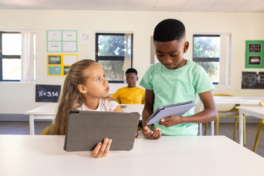 Classmates schoolchildren holding tablets in classroom, boy in green T-shirt talking to seated girl
