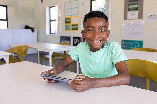African teen wearing light green tee sitting at white school table holding tablet smiling at camera