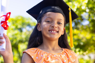 Smiling African American child in orange polka-dot dress, yellow-tassel cap holding diploma in park