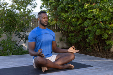 African American man in thirties meditating on yoga mat on wooden deck wearing blue athletic shirt
