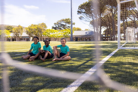 Diverse female children sitting on turf field near goal and net, wearing teal shirts barefoot