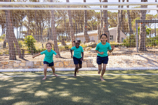 Diverse female children running from soccer goal on turf, wearing matching teal shirts