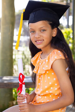 African American child sitting on bench wearing orange dress and cap with yellow tassel holding dipl