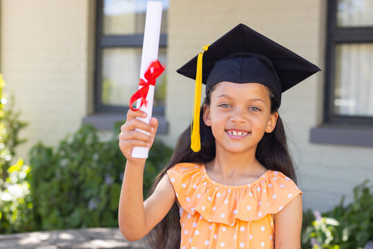 African American child wearing mortarboard and orange polkadot dress holding diploma in front yard