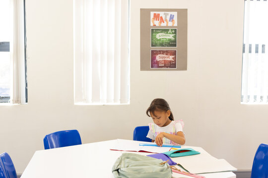 Asian child student sitting at hexagonal table in tutoring room organizing workbook, copy space