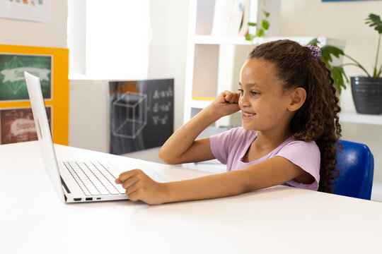 African American girl sitting at table using silver laptop, looking toward screen, hand near ear