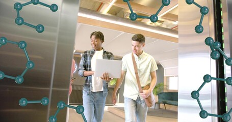 Walking two jeans-clad peers viewing laptop in office lobby with messenger bag and metal panels