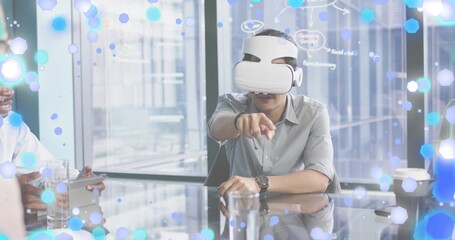Pointing man wearing gray shirt and white VR headset leaning at glass meeting table with tablet