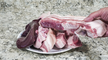 A hand lifts a cut of meat from a plate filled with various other meat cuts in a kitchen. The setting shows a countertop with stone patterns.