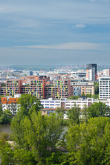 Obraz premium Urban landscape featuring colorful residential buildings alongside a river with lush green trees under a partly cloudy sky in a vibrant city setting with distant hills