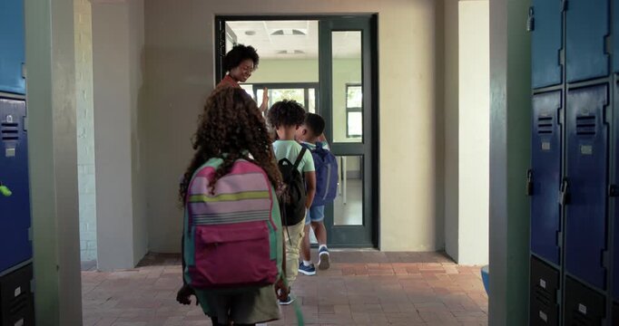 African American teacher welcoming kids entering hall, guiding backpacks through door to classroom