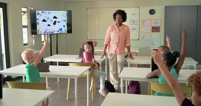 African American woman teacher in peach blouse responding to pupils raising hands in classroom