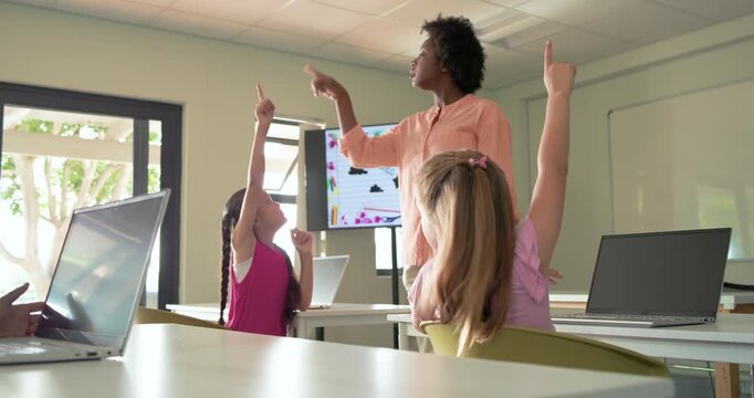 Teacher with teen girls in class asking, pointing up, girls raising hands to answer on laptops