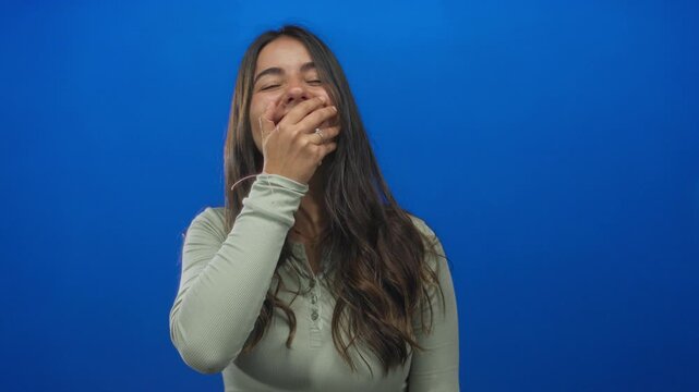 Young hispanic woman smiling and covering mouth playfully in front of an isolated blue wall background suggesting lightheartedness and fun