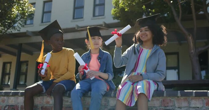 Diverse children wearing caps on low wall, right child checking then raising diploma, celebrating