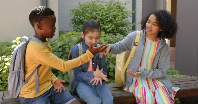 Diverse students playing hand game boy extending hand girl counting passing time on bench with bags