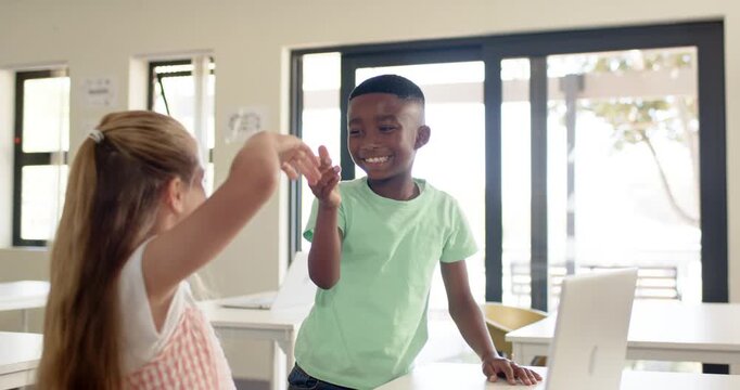 Diverse child peers in class high-fiving, boy raising hand, walking back, girl typing laptop