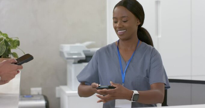 African American woman in scrubs handing phones, visitor extending phone for card reader at desk