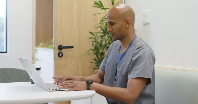 Indian man in scrubs typing on white laptop in staff room, documenting patient notes after visit