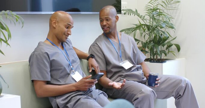 African American male staff showing phone on break in clinic lounge in scrubs and holding cup