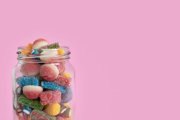 Cropped view of the upper half of a glass jar filled with colorful sugared gummy candies on the left side of a bright pink background with copy space on the right. Close-up, isolated, front view