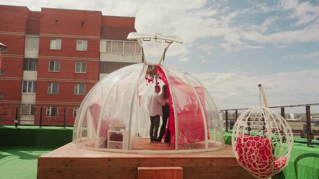 Rooftop bubble dome couple celebrating date, candlelit dinner inside transparent igloo, wooden platform, decorative flowers and string lights, urban apartment buildings in background, bright sky,