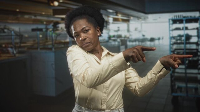 Woman points fingers emphatically to the right inside a busy commercial kitchen in a building with metal racks, trays and carts visible; determination.