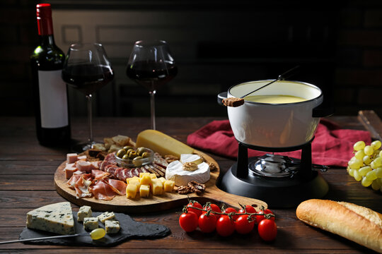 Delicious melted cheese in fondue pot, snacks and wine on wooden table against dark background, closeup