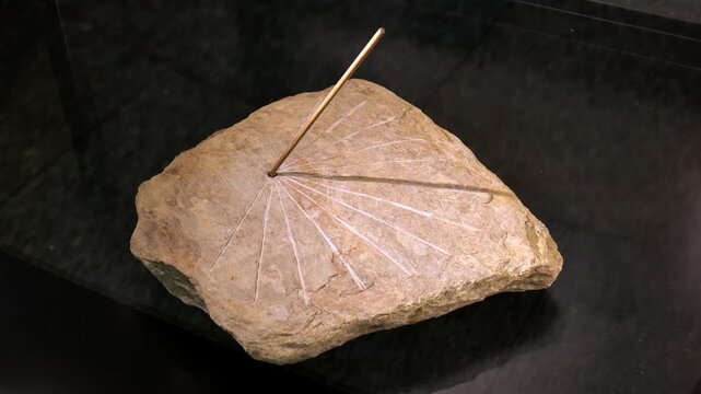 An old stone sundial with metal gnomon casting sharp shadow on rough rock slab, sunbeam rays on dark background, simple antique timekeeping instrument, history and measurement concept, no people