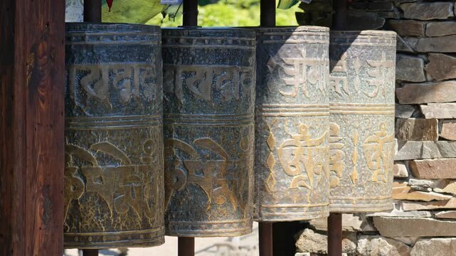 Bronze Tibetan prayer wheels with carved mantra script spinning in a row outdoors beside stone wall, sunny daylight, Buddhist monastery culture and spirituality, closeup panning shot, no people