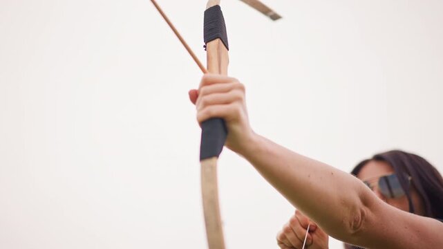 white woman nocking arrow and aiming with steady grip, closeup focus on bow and arrow against pale sky, competitive training atmosphere, sunglasses and composed posture, techniquedriven shot