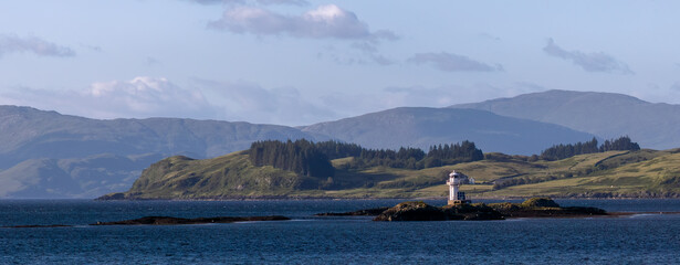 Large white lighted navigational marker sits atop a rocky outcrop in the waters of Scotland with rolling farming hills in the background. © Dawn