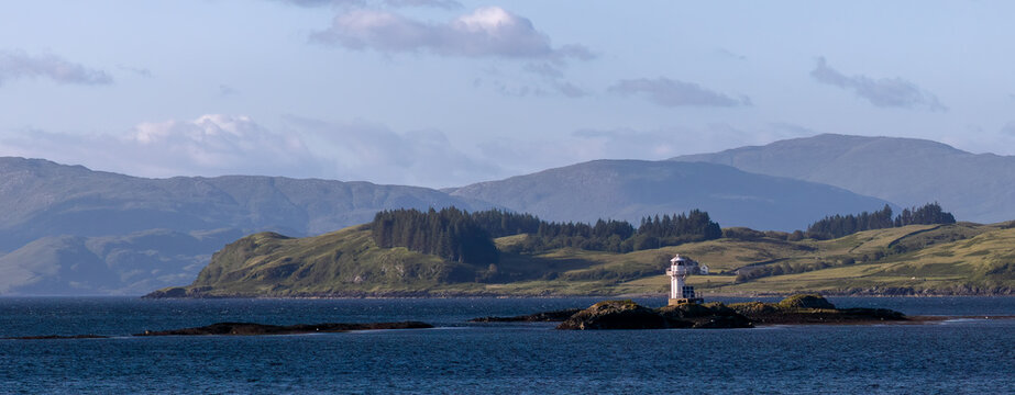 Large white lighted navigational marker sits atop a rocky outcrop in the waters of Scotland with rolling farming hills in the background.