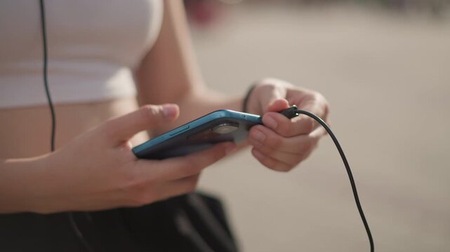 closeup hands holding phone with earphones. young woman in crop top and joggers taps screen to select playlist, adjusts wired headphone cable, sensing beat and rhythm while preparing street dance